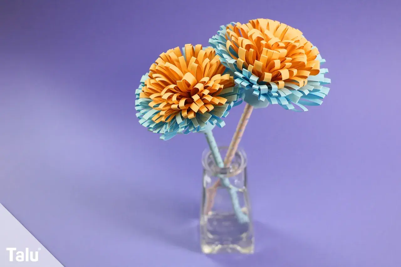 Zwei selbstgebastelte Papierblumen in Orange und Hellblau stehen in einer kleinen Glasvase. Ein schönes Beispiel für blumenstrauß basteln.