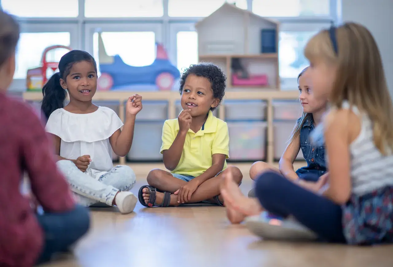 Kinder sitzen im Kreis und lernen. Die Lehrerin beobachtet aufmerksam, um die **Schulreife erkennen** zu können.