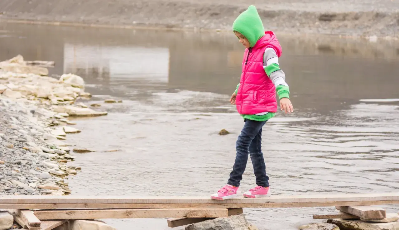 Ein Kind balanciert über ein provisorisches Holzbrett über einem Fluss. Das zeigt, was bedeutet selbstwirksamkeit: Mut, Vertrauen in die eigenen Fähigkeiten und das Überwinden von Hindernissen.