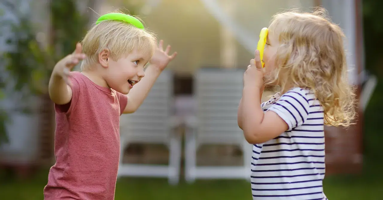 Zwei Kinder spielen im Garten. Der Junge mit einem grünen Ring auf dem Kopf lacht, das Mädchen hält ein gelbes Spielzeug. Ein Beispiel für gelebte sozialkompetenz definition.