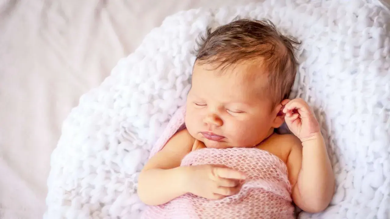 Ein schlafendes Baby, 9 Wochen alt, eingewickelt in eine rosafarbene Decke, liegt auf einer weißen, flauschigen Unterlage.