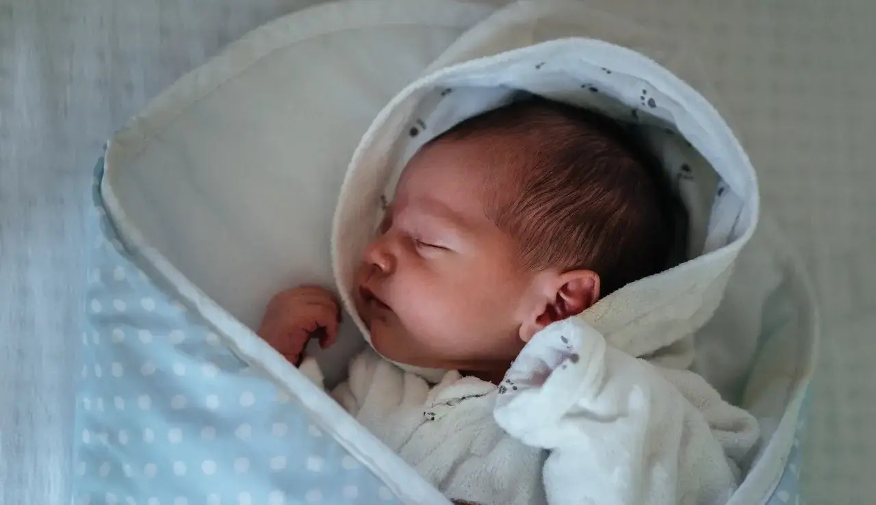 Ein friedlich schlafendes Baby, etwa 9 Wochen alt, eingewickelt in eine weiche Decke mit blauen Punkten.