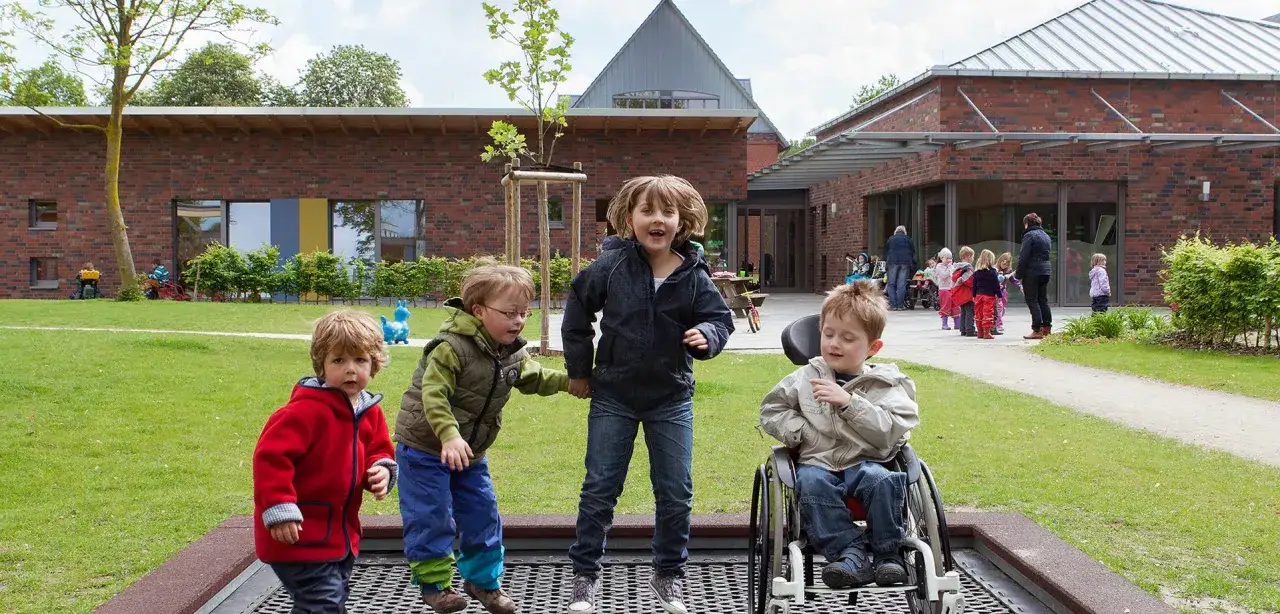 Kinder mit und ohne Autismus spielen gemeinsam im Kindergarten. Ein Junge im Rollstuhl lacht, w&auml;hrend andere Kinder auf einem Trampolin springen.