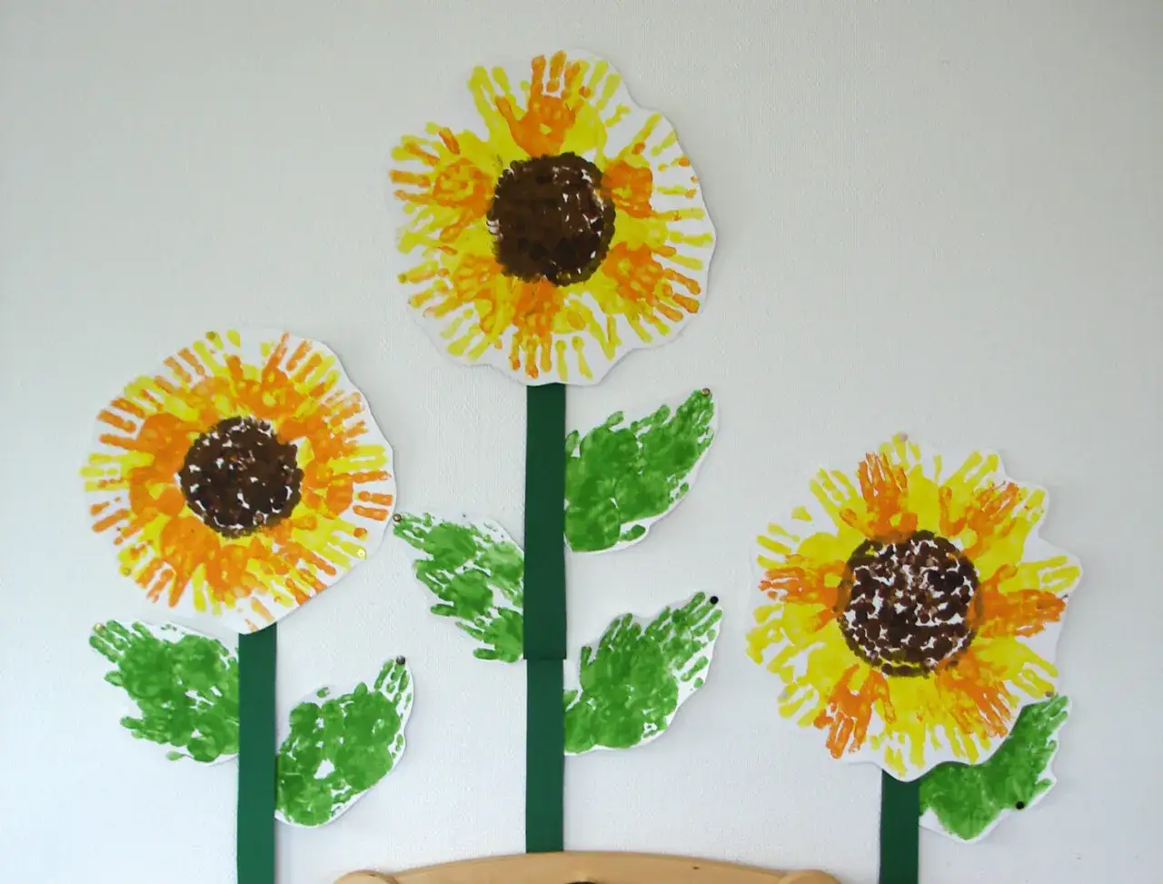 Sonnenblume basteln Krippe: Drei bunte Sonnenblumen aus Handabdrücken, mit grünen Blättern und Stielen, an einer weißen Wand.