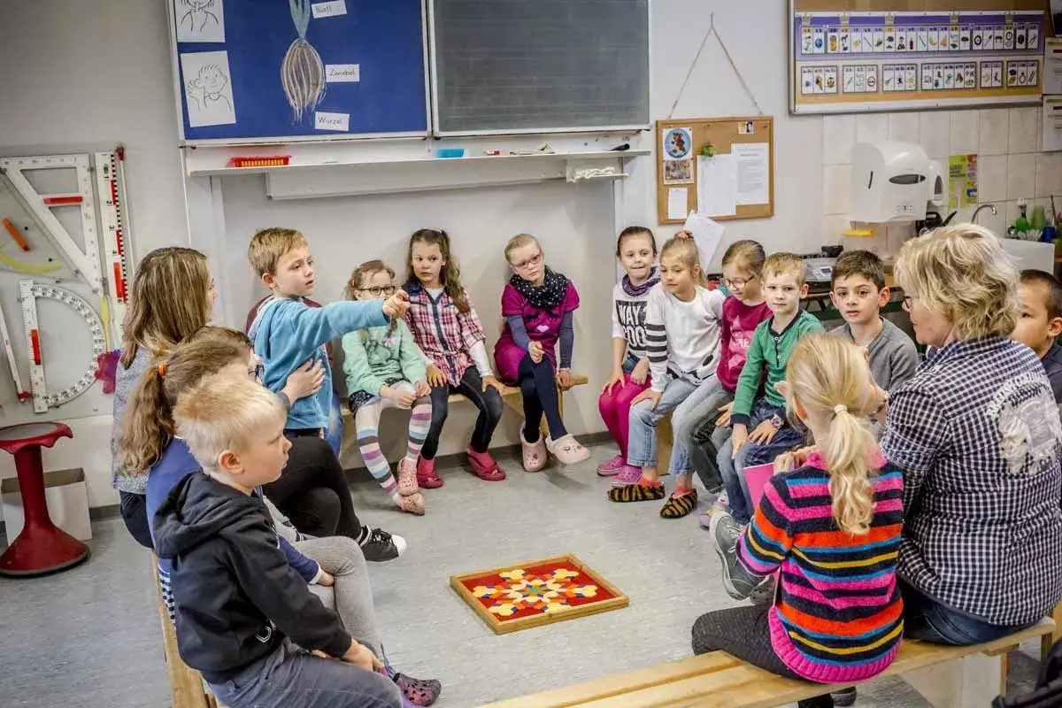Kinder und Lehrerin im Kreis sitzend, ein Junge zeigt auf ein Spielbrett. Dies ist ein Beispiel für die eingangsdiagnostik grundschule.