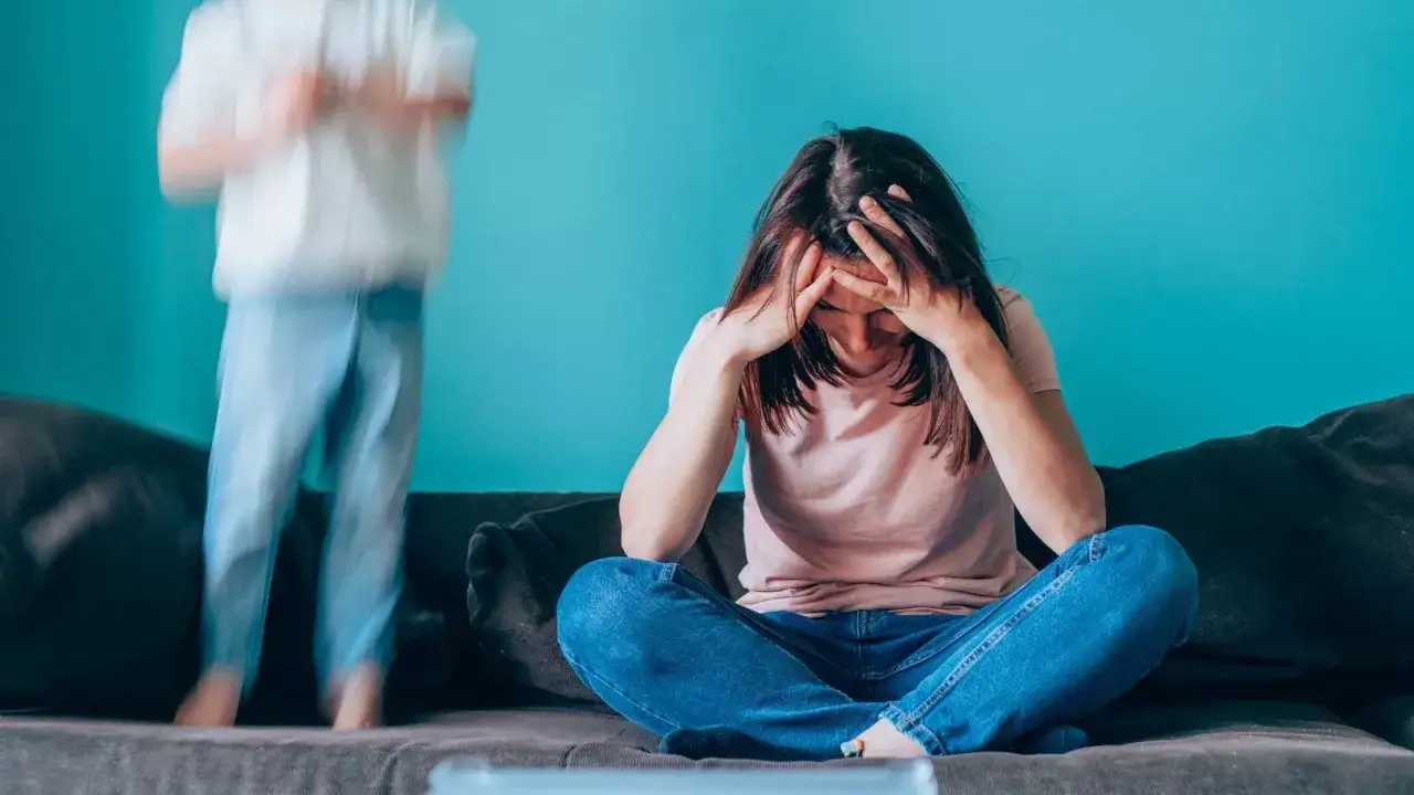 Erschöpfte Mutter mit Händen am Kopf, während ein Kind im Hintergrund herumtollt. Ein Bild von Mama Burnout.