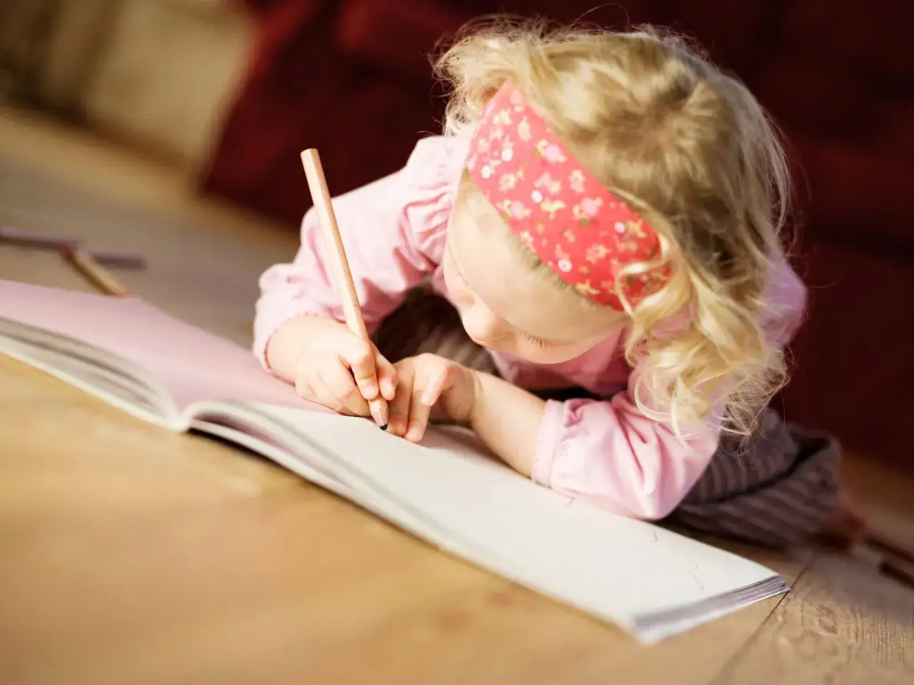 Kleines Mädchen mit Bleistift auf dem Boden, das in ein Buch schreibt. Mögliche Ursachen für schlechte Handschrift bei Kindern sind mangelnde Feinmotorik oder Konzentration.