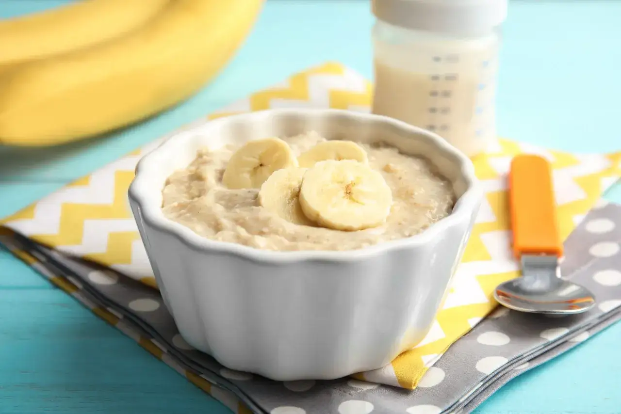 Schüssel mit Brei und Bananenscheiben, daneben eine Flasche mit baby kuhmilch. Ein Löffel liegt auf einer Serviette.