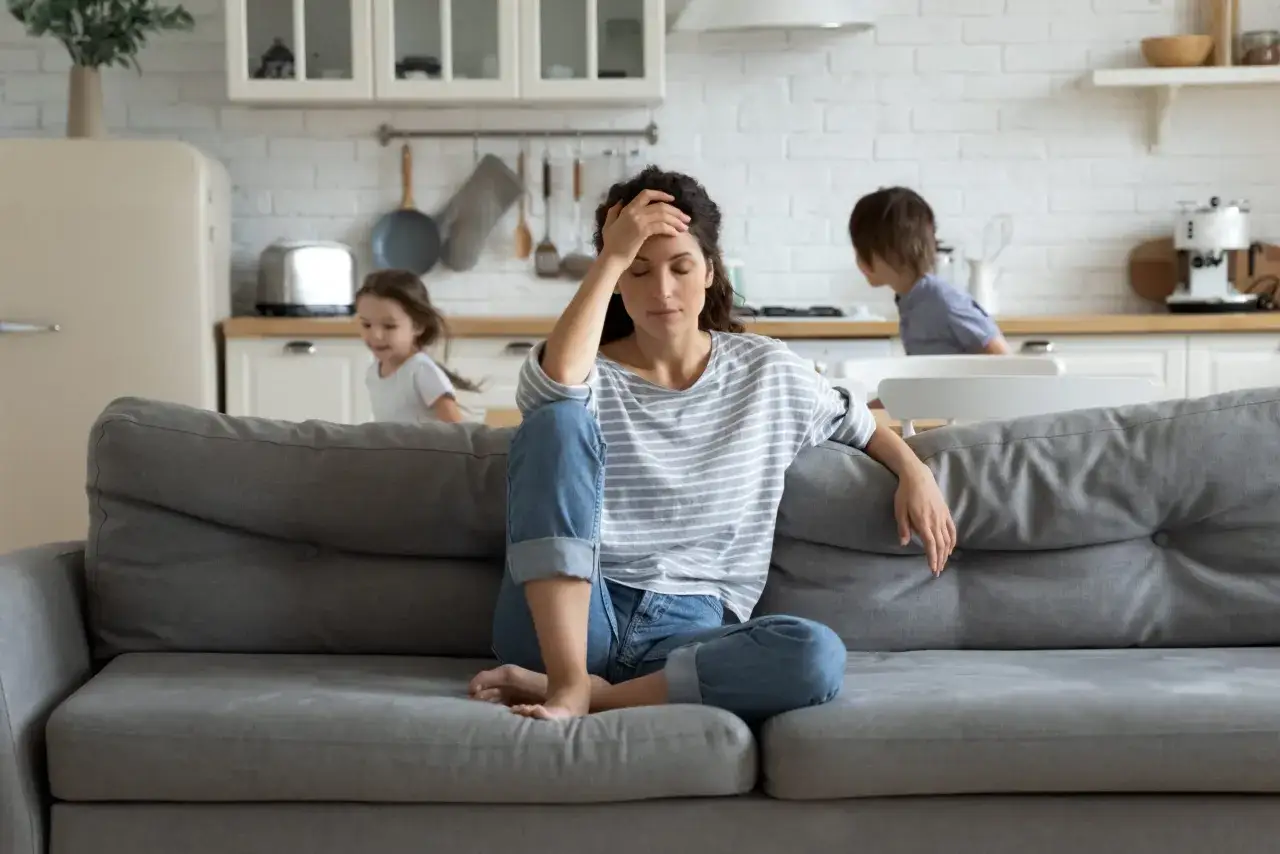 Frau sitzt erschöpft auf dem Sofa, Kinder spielen im Hintergrund. Deutliche Anzeichen von emotionaler Stress Symptome.