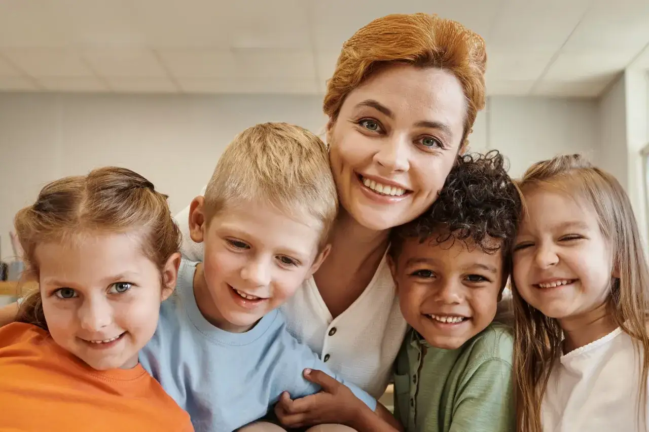 Eine lächelnde Erzieherin umarmt vier fröhliche Kinder unterschiedlicher Herkunft. Ein Beispiel für gelebte Inklusion in der Kita.