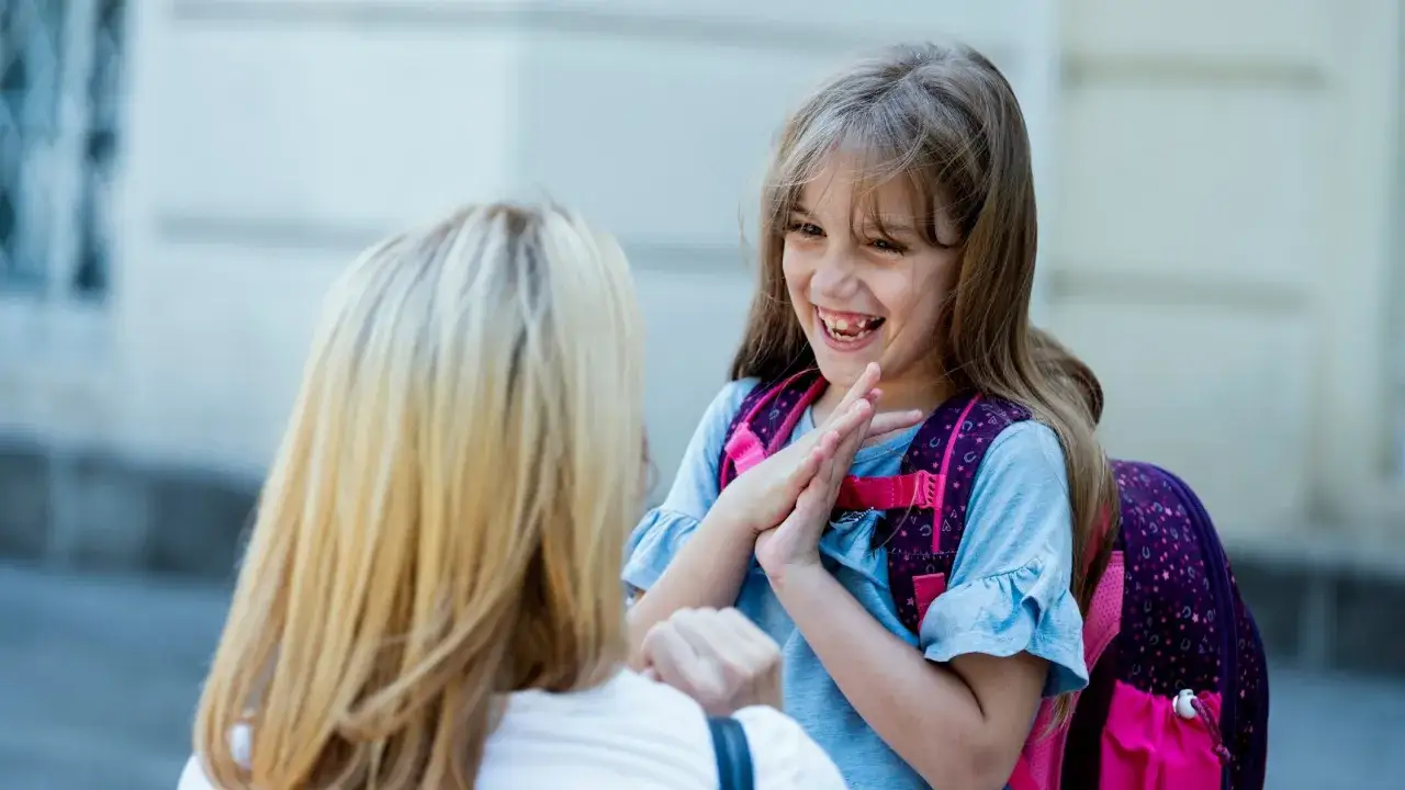 Ein lachendes 7-jähriges Mädchen mit Rucksack gibt einer Frau High-Five. Trotz einer schwierigen Phase strahlt sie Freude aus.