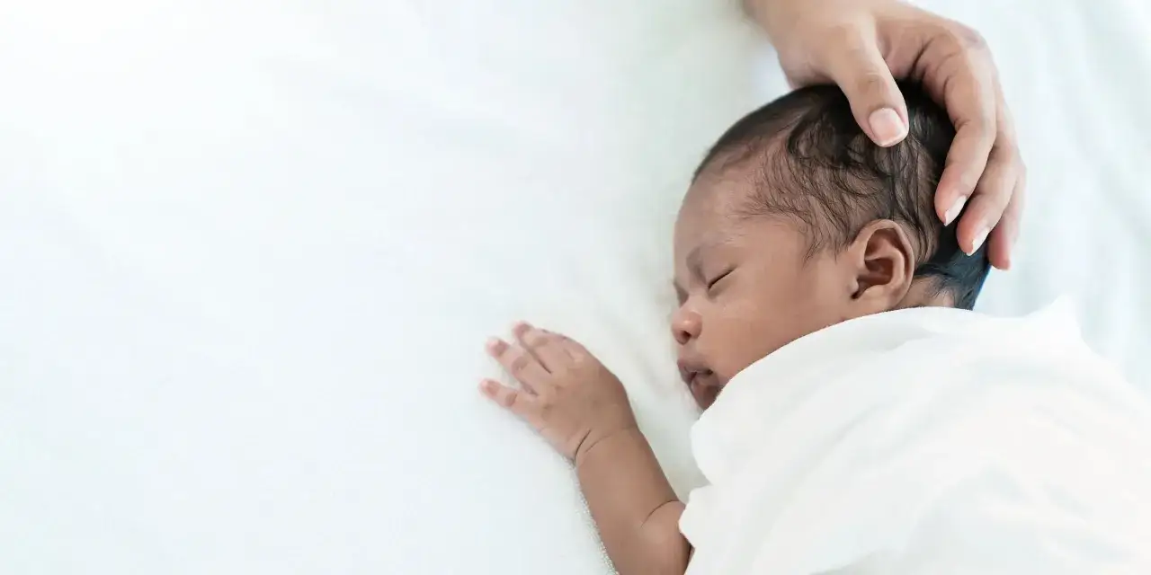 Ein s&uuml;&szlig;es Baby schwitzt beim Schlafen am Kopf, w&auml;hrend eine Hand sanft den Kopf streichelt.