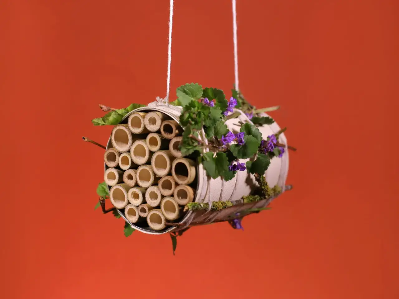 Ein selbstgebautes Insektenhotel f&uuml;r Kinder, gef&uuml;llt mit Bambusr&ouml;hren und verziert mit gr&uuml;nen Bl&auml;ttern und lila Bl&uuml;ten.