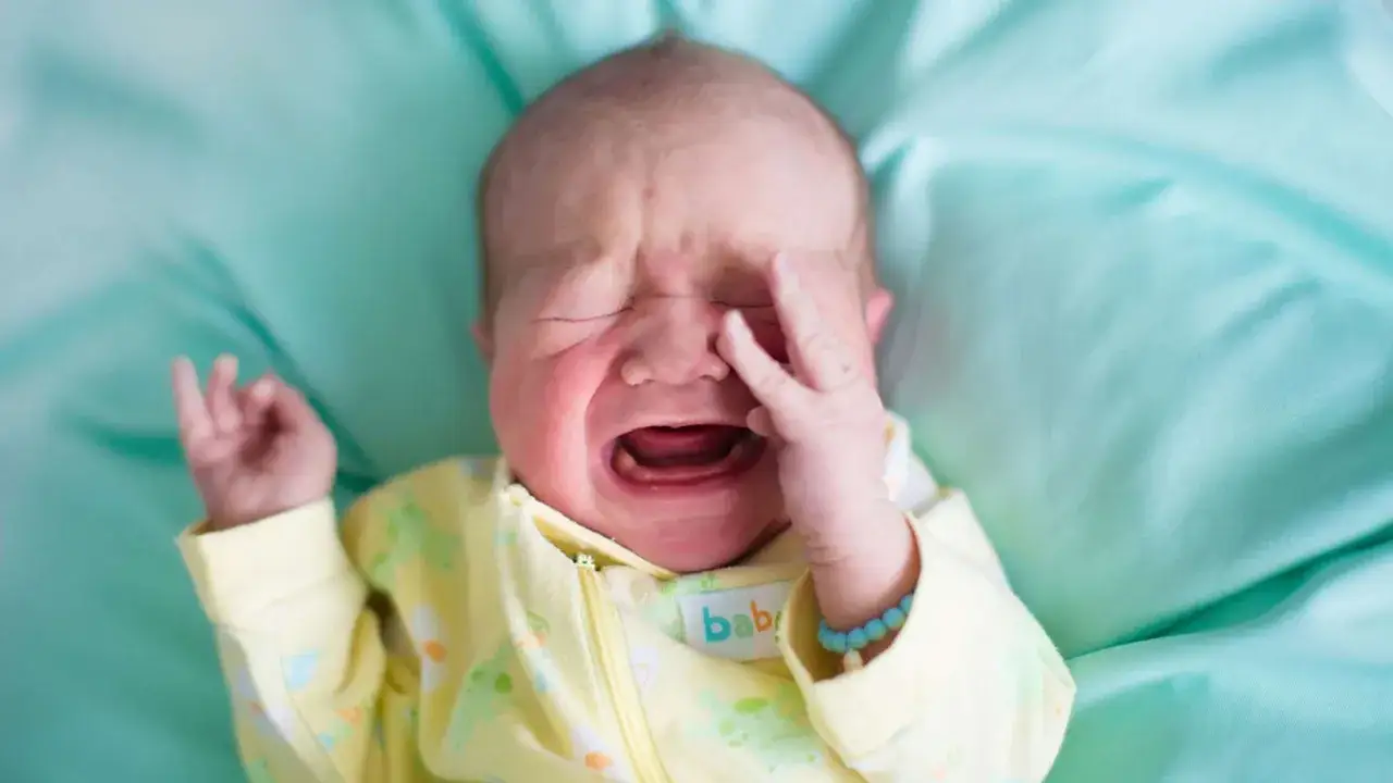 Ein weinendes Baby in gelbem Strampler, das sich mit der Hand das Auge reibt. Die 