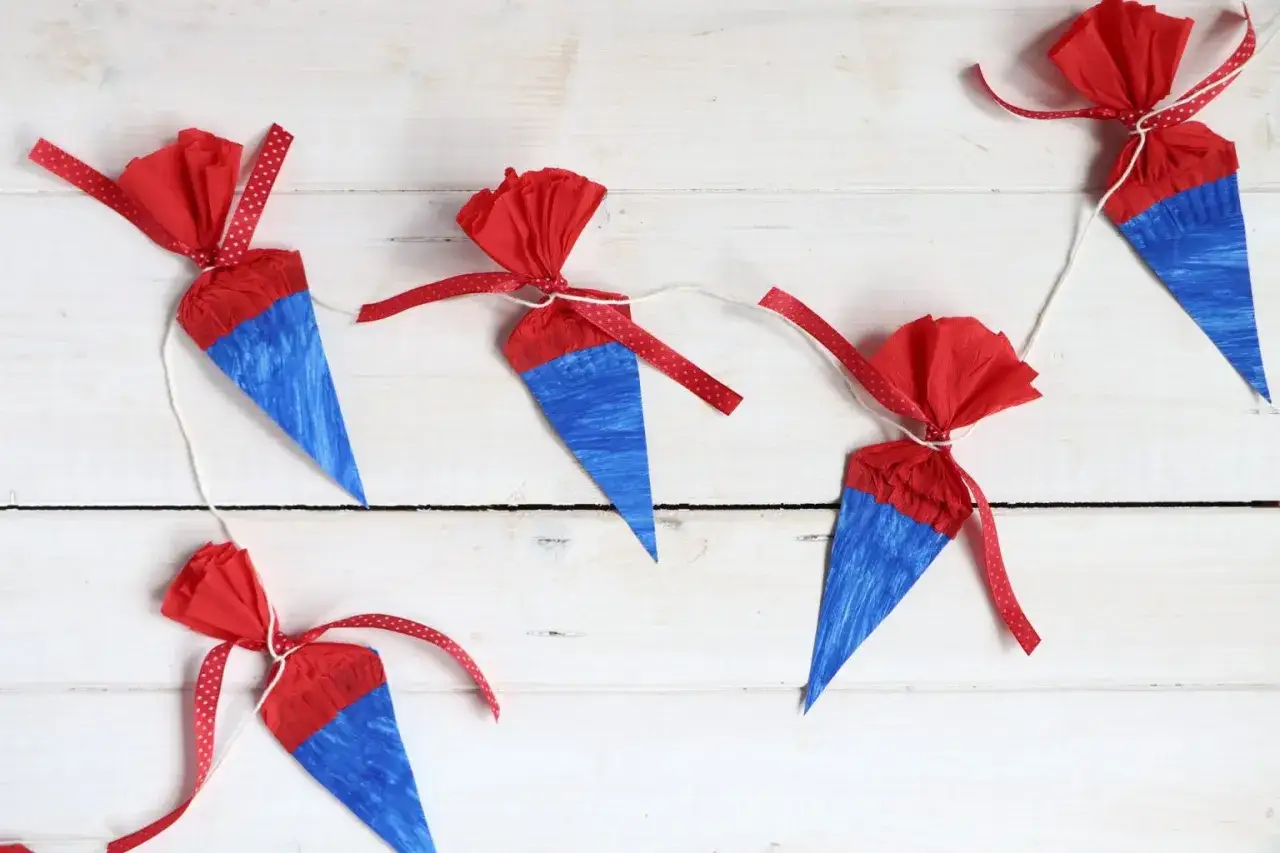 Bunte Schult&uuml;ten-Girlande als Deko-Ideen f&uuml;r die Einschulung. Rote Papierbl&uuml;ten und blaue Kegel mit roten B&auml;ndern.