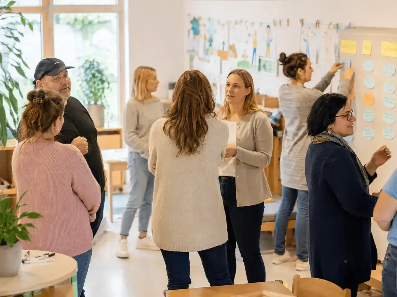 Eltern diskutieren Ideen für den Elternabend in der Krippe. Eine Frau heftet Notizen an eine Wand, während andere sich unterhalten.