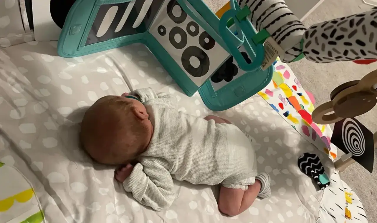 Ein baby 10 wochen alt schläft auf einer Spieldecke. Bunte Spielzeuge sind um das Baby herum angeordnet.