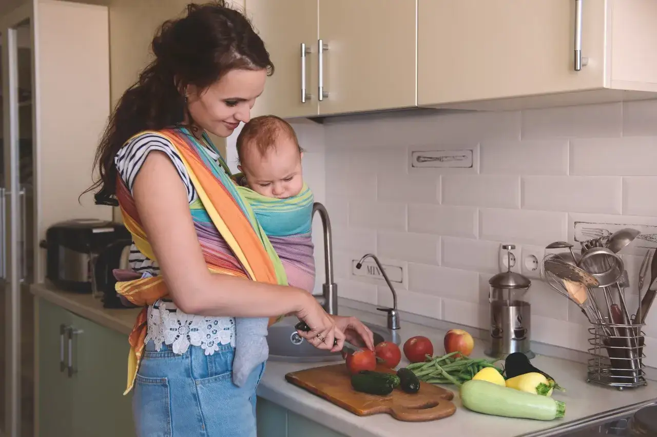 Mutter bereitet scharfes Essen zu, während sie ihr Baby stillt. Sie schneidet Gemüse in der Küche.