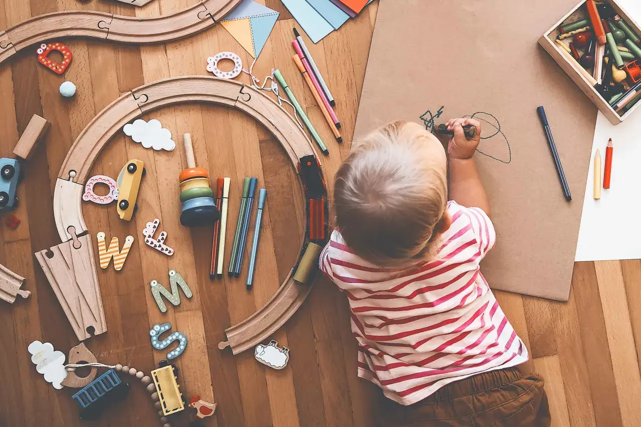 Kind malt mit Stiften, umgeben von Holzspielzeug wie einer Eisenbahn und Buchstaben. Das ist, was Montessori ausmacht: freies Spiel und Lernen.