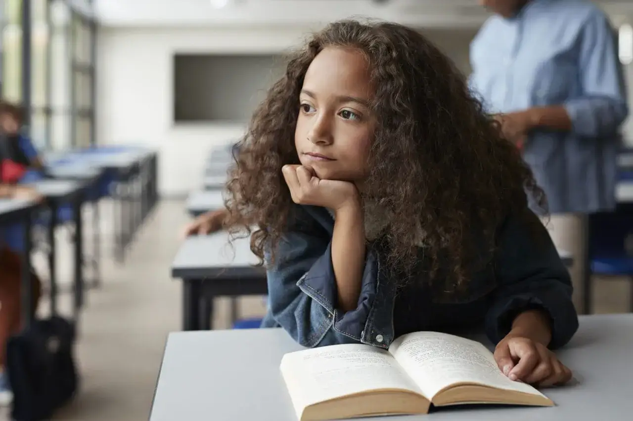 Ein Mädchen mit Locken sitzt nachdenklich im Klassenzimmer, die Hand am Kinn. Der Blick ist nach außen gerichtet, was auf Stress bei Kindern hindeuten könnte.
