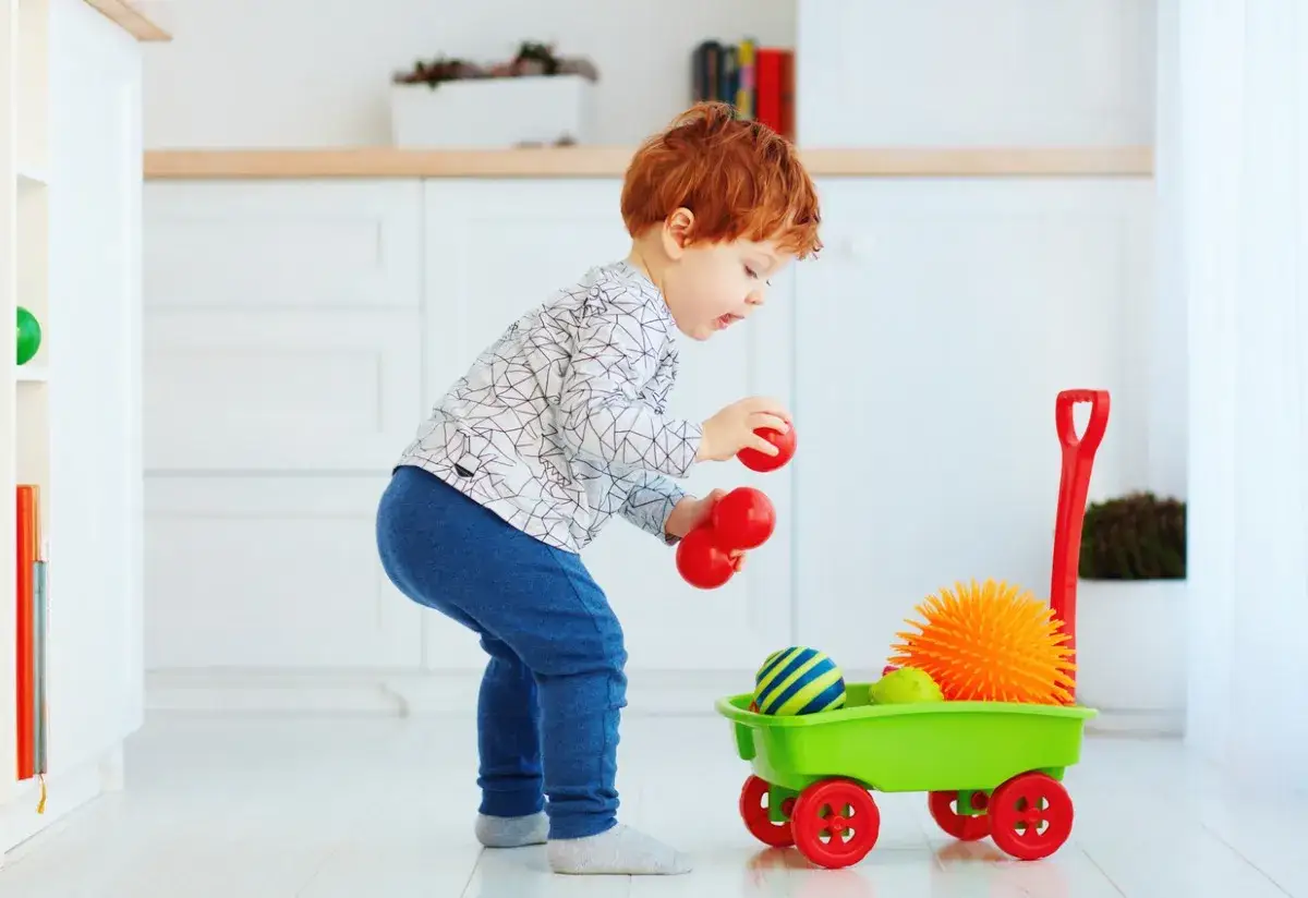 Rotschopf-Kleinkind mit roten Bällen in der Hand, spielt mit einem grünen Spielzeugwagen voller bunter Bälle.