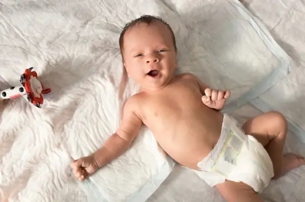 Ein fröhliches Baby, etwa 6 Wochen alt, liegt auf einer weißen Unterlage und blickt neugierig in die Kamera. Neben ihm liegt ein buntes Spielzeug.