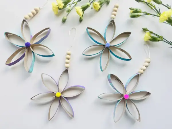 Bunte Blumen aus Klopapierrollen basteln: Vier hübsche Anhänger mit Holzperlen und Pompons, umgeben von gelben Blüten.