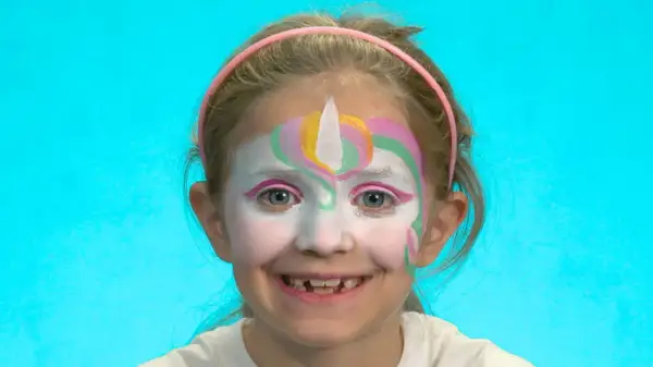 Ein Mädchen mit einem bunten Einhorn-Schminken für Kinder. Das Gesicht ist weiß grundiert, mit einem weißen Horn und bunten Bögen in Rosa, Gelb und Grün.