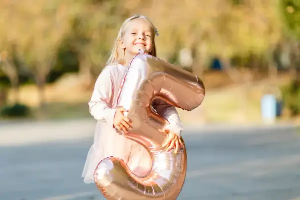 Ein glückliches, blondes Kind von 5 Jahren hält stolz einen großen, roségoldenen Ballon in Form der Zahl 5.