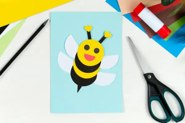 Süße Biene basteln: Gelber Kopf mit Herzfühler, schwarze Streifen, weiße Flügel auf hellblauem Papier. Schere und Klebestift liegen bereit.