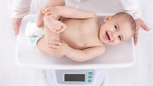 Ein lächelndes Baby liegt auf einer Waage für die U3 Untersuchung. Hände halten das Kind sanft.