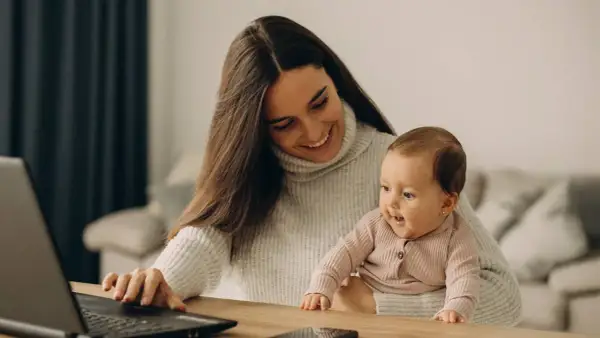 Lächelnde Mutter mit Baby auf dem Schoß am Laptop. Ein Bild, das den Spagat zwischen Beruf und Familie zeigt, oft verbunden mit dem Gefühl von Mama Burnout.