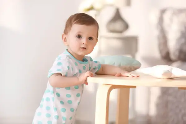 Ein neugieriges Baby, etwa 1 Jahr alt, stützt sich auf einen Holztisch. Es trägt ein weißes T-Shirt mit blauen Tupfen.