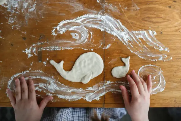 Kinderhände gestalten Salzteig-Ideen: zwei Wale schwimmen in Meereswellen aus Mehl auf einem Holztisch.