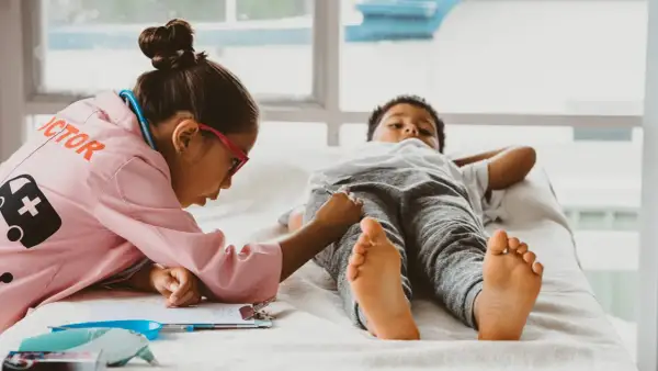 Kinder spielen Doktorspiele im Kindergarten. Ein Mädchen in Arztkittel untersucht einen Jungen, der auf einer Liege liegt.