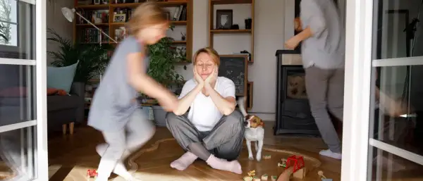 Chaos im Wohnzimmer: Kinder rennen herum, ein Hund schaut neugierig, und die mama steht kopf. Sie sitzt auf dem Boden, die Augen geschlossen, während das Leben um sie herum tobt.