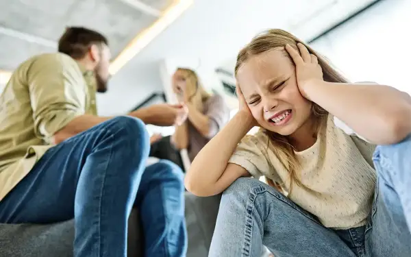 Ein Kind hält sich die Ohren zu, während Eltern im Hintergrund streiten. Das aggressive Verhalten der Erwachsenen belastet das Kind sichtlich.