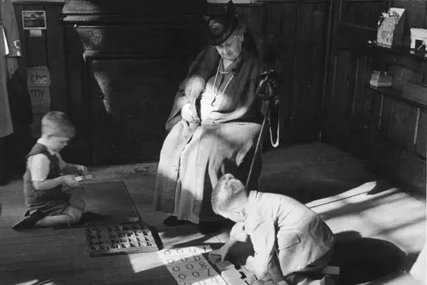 Maria Montessori beobachtet Kinder beim Lernen. Ein Junge kniet und sortiert Buchstaben, ein anderer übt mit Zahlen.