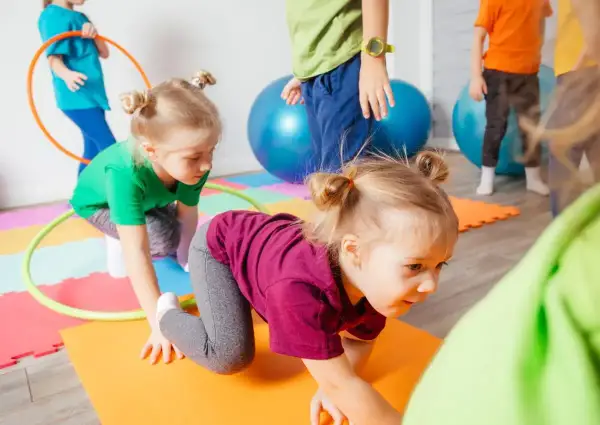 Kinder haben Spaß bei Bewegungsspielen im Kindergarten. Sie krabbeln auf bunten Matten, umgeben von Gymnastikbällen und Reifen.