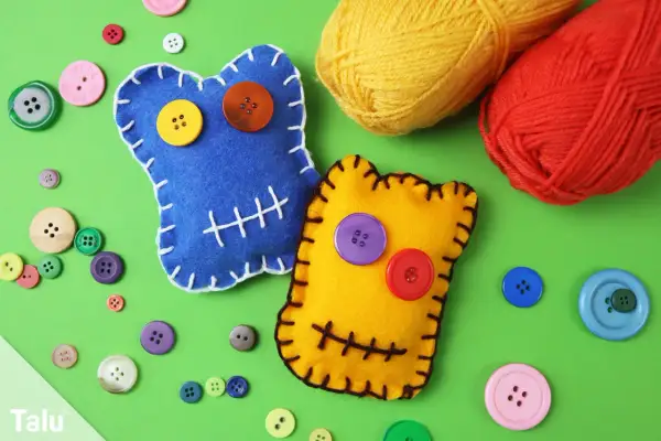Zwei bunte Filz-Wutmonster basteln: Ein blaues mit gelbem und orangefarbenem Knopfauge, ein gelbes mit lila und rotem Knopfauge. Viele bunte Knöpfe und Garnknäuel liegen daneben.
