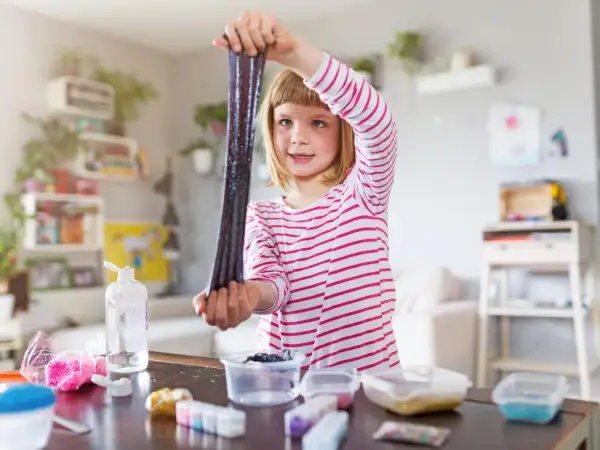 Ein Mädchen hält glitzernden, dunklen Schleim hoch, den sie gerade selber gemacht hat. Auf dem Tisch liegen Zutaten für weitere Schleim-Kreationen.