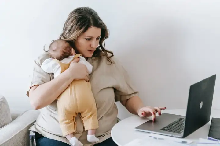 Alleinerziehend und im **burnout**: Eine Frau hält ihr Baby und arbeitet am Laptop.